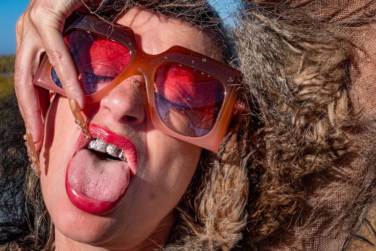 Close-up portrait of Canadian musician Peaches wearing sunglasses with red tinted lenses, bright lipstick, and a diamond grillz tooth gem.